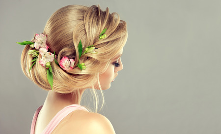 Young, attractive woman is demonstrating blonde hair gathered in elegant evening or wedding hairstyle with fresh flowers in it.  Hairdressing art, coloration of hair and beauty products. View from back side.の写真素材