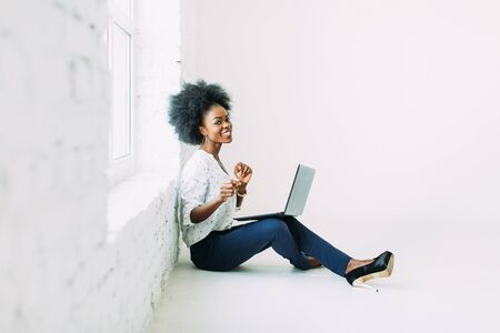 Young african american business woman using the laptop, while sitting on the floor near a big window in studioの写真素材