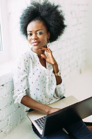 Young smiling african american business woman using the laptop, sitting on the floor in white light studioの写真素材
