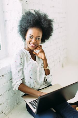 Young smiling african american business woman using the laptop, sitting on the floor in white light studioの写真素材