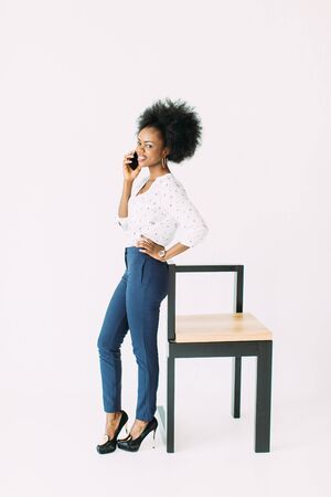 cheerful young afro american business woman talking on cell phone, standing near the modern chair, isolated on whiteの写真素材
