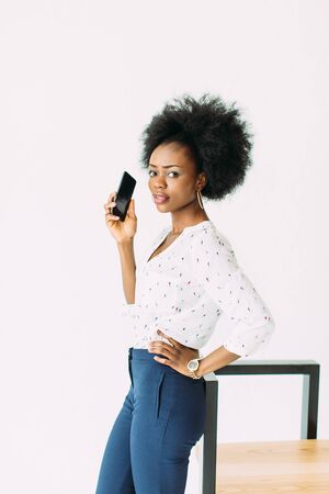 cheerful young afro american business woman talking on cell phone, standing near the modern chair, isolated on whiteの写真素材