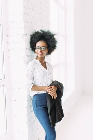 Young african american business woman in glasses, standing near the big window and holding jacketの写真素材