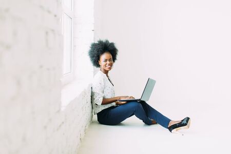 Young african american business woman using the laptop, while sitting on the floor near a big window in studioの写真素材