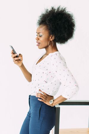 cheerful young afro american business woman talking on cell phone, standing near the modern chair, isolated on whiteの写真素材