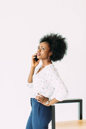cheerful young afro american business woman talking on cell phone, standing near the modern chair, isolated on whiteの写真素材