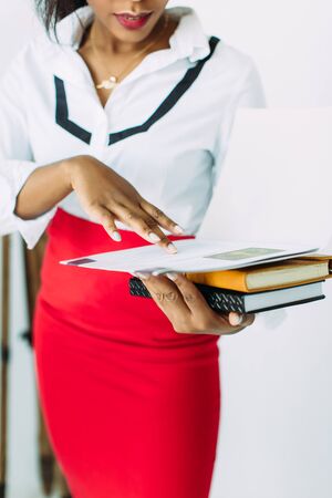Beautiful young african smiling woman teacher in white shirt and red skirt holding books and documentsの写真素材