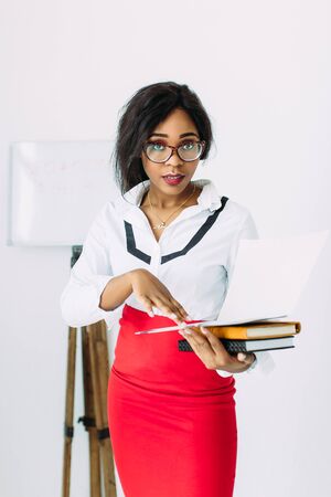 Beautiful young african smiling woman teacher in white shirt and red skirt holding books and documentsの写真素材