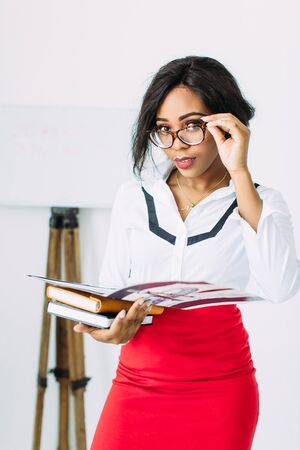 Beautiful young african smiling woman teacher in white shirt and red skirt holding books and documentsの写真素材