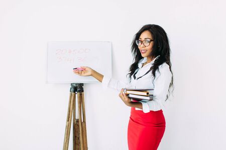 Beautiful young african smiling woman teacher holding documents, books, standing near the boardの写真素材