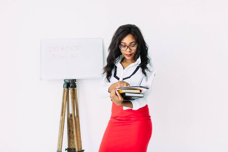 Beautiful young african smiling woman teacher holding documents, books, standing near the boardの写真素材