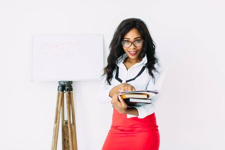 Beautiful young african smiling woman teacher holding documents, books, standing near the boardの写真素材
