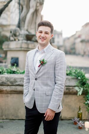 stylish smiling groom in a gray jacket, posing outdoors, standing on the stone stairs of the monument in old city center before the wedding ceremonyの写真素材