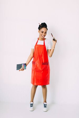 Beautiful young African American beautician woman holding set of make up brushes and eye-shadows, isolated on whiteの写真素材