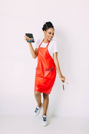 Beautiful young African American beautician woman holding set of make up brushes and eye-shadows, isolated on whiteの写真素材