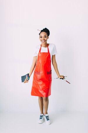 Beautiful young African American beautician woman holding set of make up brushes and eye-shadows, isolated on whiteの写真素材
