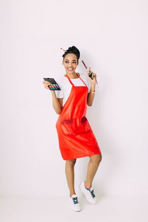 Beautiful young African American beautician woman holding set of make up brushes and eye-shadows, isolated on whiteの写真素材
