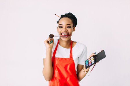 Beautiful young African American beautician woman holding set of make up brushes and eye-shadows,...