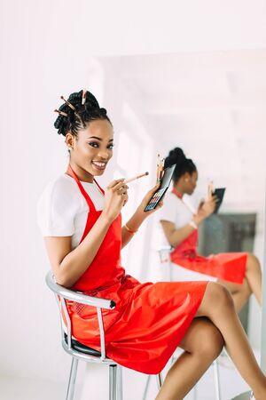 Beautiful young African American beautician woman holding set of make up brushes and watching in the mirrorの写真素材