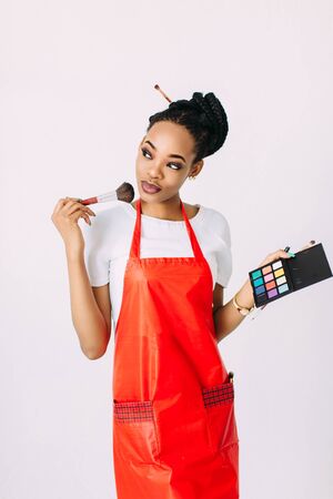 Beautiful young African American beautician woman holding set of make up brushes and eye-shadows, isolated on whiteの写真素材