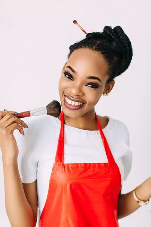 Beautiful young African American beautician woman holding set of make up brushes and eye-shadows, isolated on whiteの写真素材