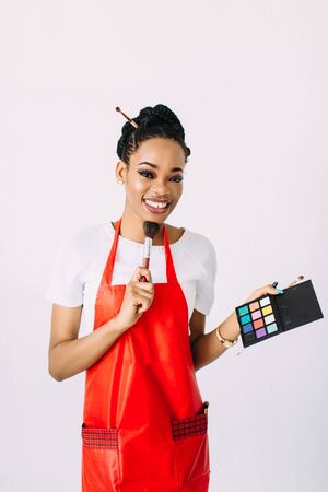 Beautiful young African American beautician woman holding set of make up brushes and eye-shadows, isolated on whiteの写真素材