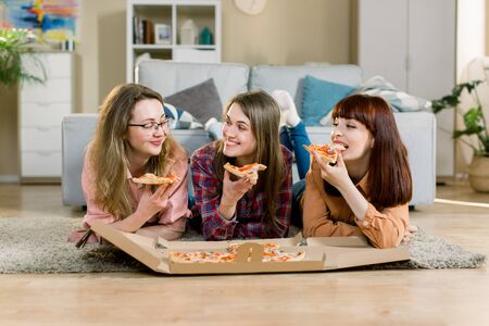 Eating Fast Food. Happy three Beautiful Friends Laughing, Eating Pizza At Home Party. Women Having Dinner Together, Enjoying Meal. Leisure, Friendship, Celebration And People Conceptの写真素材