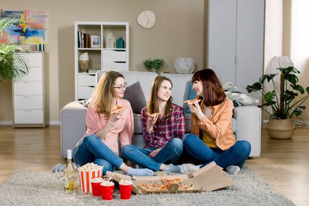 friendship, holidays, fast food and celebration concept - happy young women friends with drinks and popcorn eating pizza at home, sitting on the floor in cozy roomの写真素材