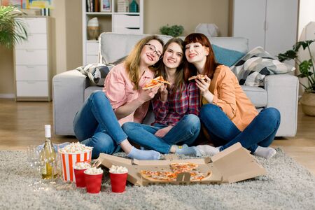 friendship, holidays, fast food and celebration concept - happy young women friends with drinks and popcorn eating pizza at home, sitting on the floor in cozy roomの写真素材