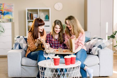 Three women friends having dinner at home. Photo of cheerful surprised girls celebrating a party at home, opening cardboard box with with pizza.の写真素材