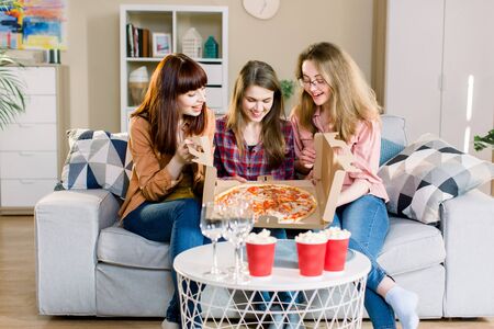 Three women friends having dinner at home. Photo of cheerful surprised girls celebrating a party at home, opening cardboard box with with pizza.の写真素材