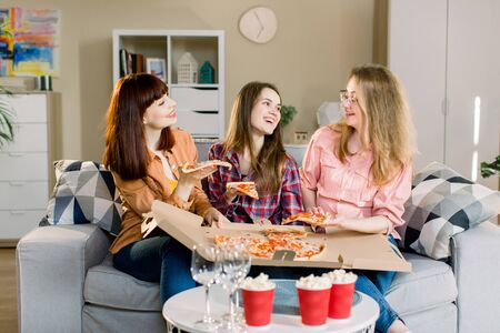 Portrait of three happy young girlfriends eating pizza on sofa at home. Beautiful ladies smiling and communicating about every day life: work, home, men.の写真素材