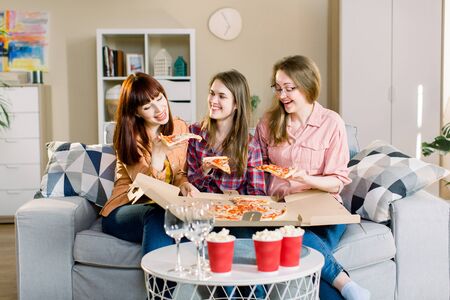 Portrait of three happy young girlfriends eating pizza on sofa at home. Beautiful ladies smiling and communicating about every day life: work, home, men.の写真素材