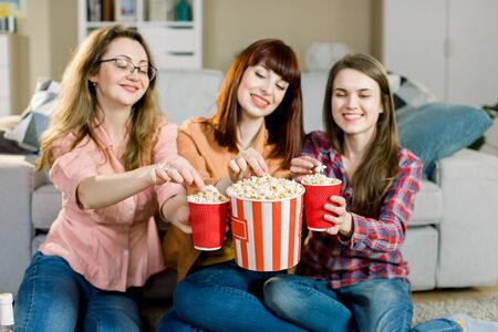 Happy three young female friends eating popcorn and sitting on the floor near sofa at homeの写真素材