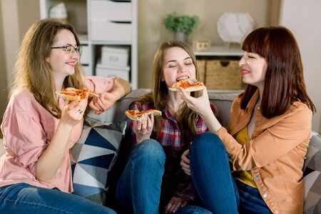 friendship, people, pajama party and junk food concept - happy young three women or girls eating pizza, popcorn and drinking wine at homeの写真素材