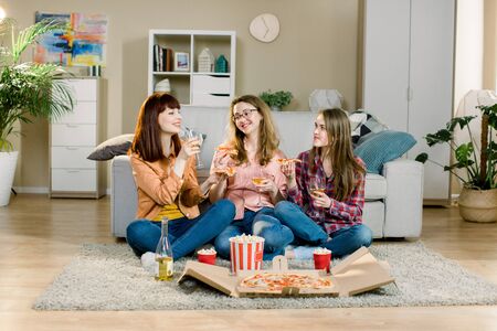 friendship, holidays, fast food and celebration concept - happy young women friends with drinks and popcorn eating pizza at home, sitting on the floor in cozy roomの写真素材