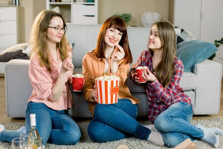 Women friendship, home party. Three beautiful funny young girl friends with popcorn in hands sitting on the floor in cozy room at home and looking on cameraの写真素材