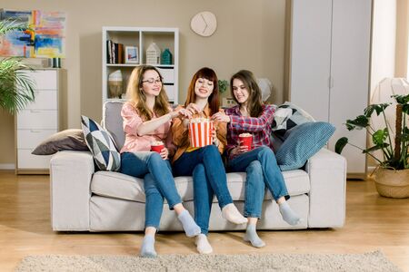 Three beautiful young women friends at home eating popcorn sitting on gray sofa together and laughing. Home women partyの写真素材