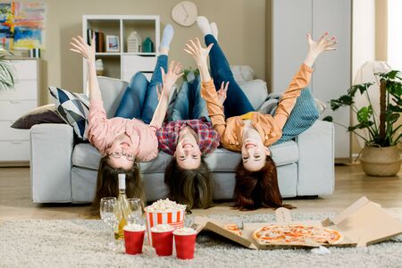 Portrait of crazy funny three women in casual outfit lying upside down on sofa holding hands up and looking at camera indoor. Home pizza partyの写真素材