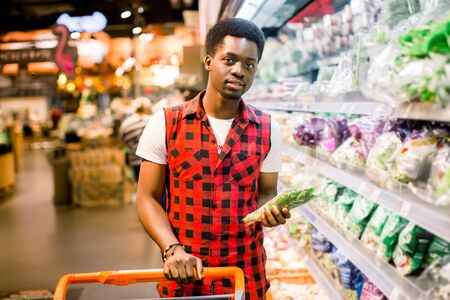 African man shopping in vegetable section at supermarket. Black man doing shopping at market while buying vegetables. Handsome guy holding shopping basket reading nutritional values of product.の写真素材