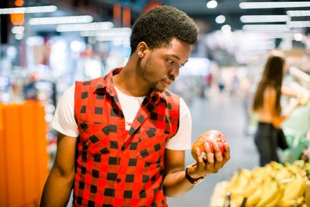 Young african man buying vegetables and fruits in grocery section at supermarket. Black man choose vegetables and fruits in the supermarket while holding grocery basket.の写真素材