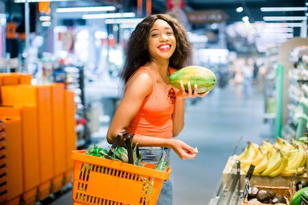 happy african american woman with shopping basket in vegetable and fruit department in supermarketの写真素材