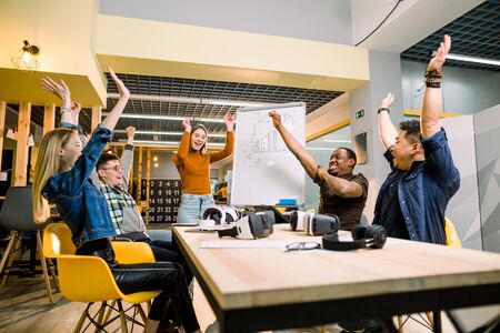 Young business team with hands up for new startup with vr goggles or victory in creative office. Cheerful diverse people giving strength motivation. Co-working and teamwork conceptの写真素材