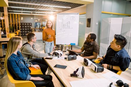 Young woman standing near the white office board with chart and speaking to her colleagues . Multiracial team of people having meeting, testing virtual reality glasses in office sitting at the tableの写真素材