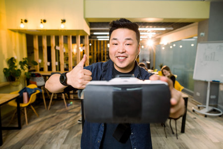 Cheerful Asian man smiling, holding virtual reality 3d glasses and thumb up, copy space. Happy male entrepreneur using 3d vr headset at workplace. Cheerful businessman with virtual reality goggles.の写真素材
