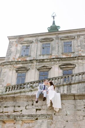 Beautiful Asian wedding couple poses while sitting on the stone wall of an old ruined castleの写真素材