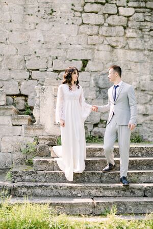 Romantic newlywed Asian couple posing in the ruins of an old castle, holding hands and standing on the stone stairsの写真素材