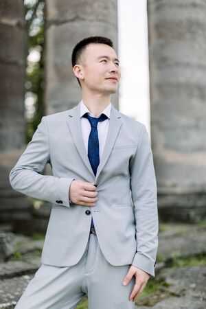 Elegant smiling handsome Asian groom in gray suit posing on the background of ancient stone columns in old castleの写真素材