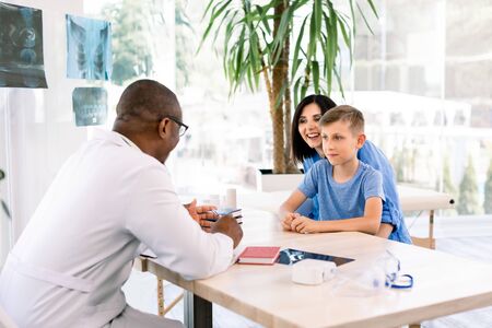 cheerful caucasian mother and her son in doctors office with african doctor, sitting at the table. Healthcare and pediatrics conceptの写真素材
