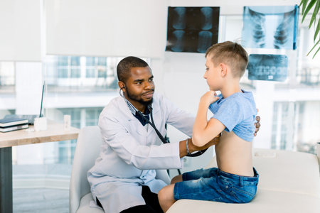 African male doctor and child boy patient. Smiling pediatrician man sees boy in medical office. African American male pediatrician with stethoscope listening to lung and heart sound of little boyの写真素材